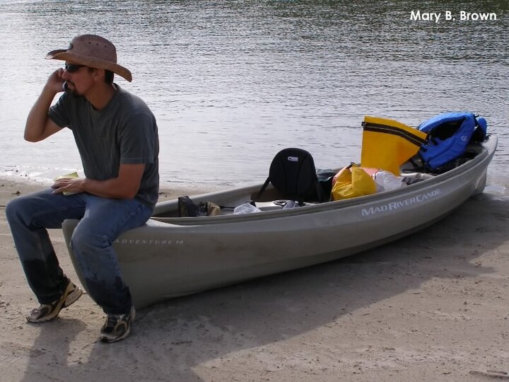 Joel Jorgensen on the phone on a canoe on a river sandbar